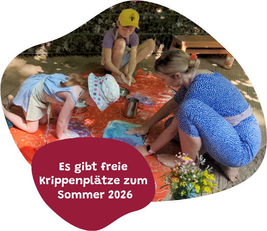 Eine Erzieherin und zwei Kindern beim Nassfilzen auf dem Boden. Sie arbeiten auf einer orangefarbenen Wachstuckdecke. Davor ist ein Feld auf dem steht: Freie Krippenplätze zum Sommer 2026